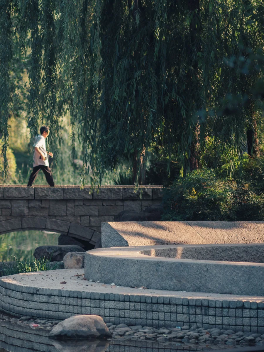 A solo visitor crossing a stone footbridge through a green park.